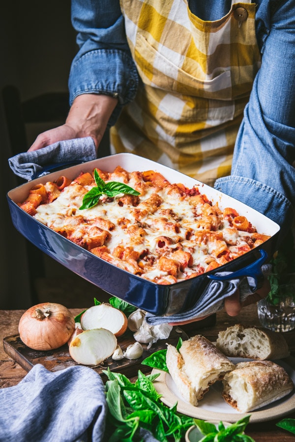 Side shot of hands holding a blue casserole dish full of baked rigatoni and cheese with fresh herbs on top