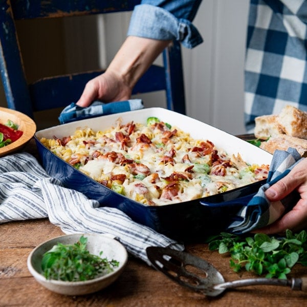 Square shot of hands holding a pan of chicken broccoli bacon ranch casserole on a wooden table