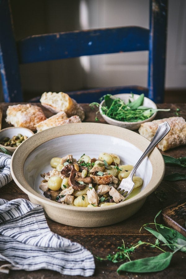 Side shot of a bowl of gnocchi on a table with a side of bread and fresh herbs