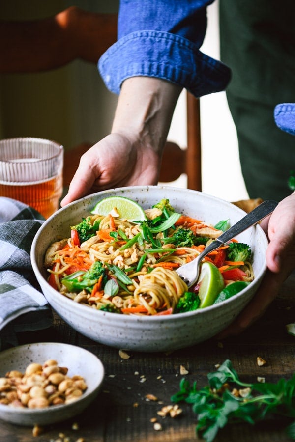 Hands serving a white bowl of easy chicken lo mein recipe on a wooden table