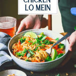 Hands serving a bowl of easy chicken lo mein on a wooden table with text title overlay