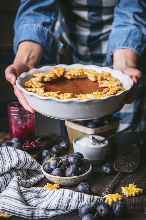 Hands holding a damson plum pie in front of a blue and white apron.