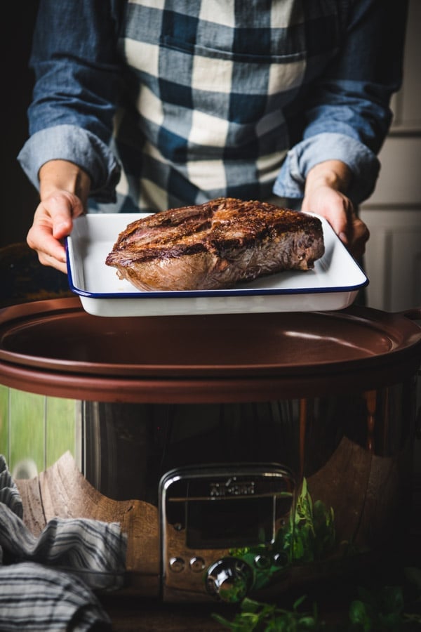 A woman holds a seared chuck roast on a pan over a large open crockpot, ready to make Italian beef sandwiches.