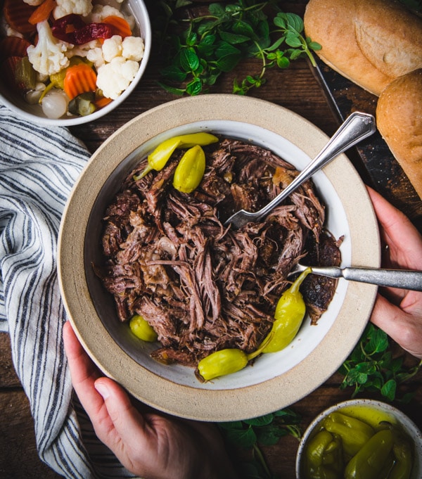 Overhead shot of hands holding a bowl of slow cooker Italian beef with two forks for shredding
