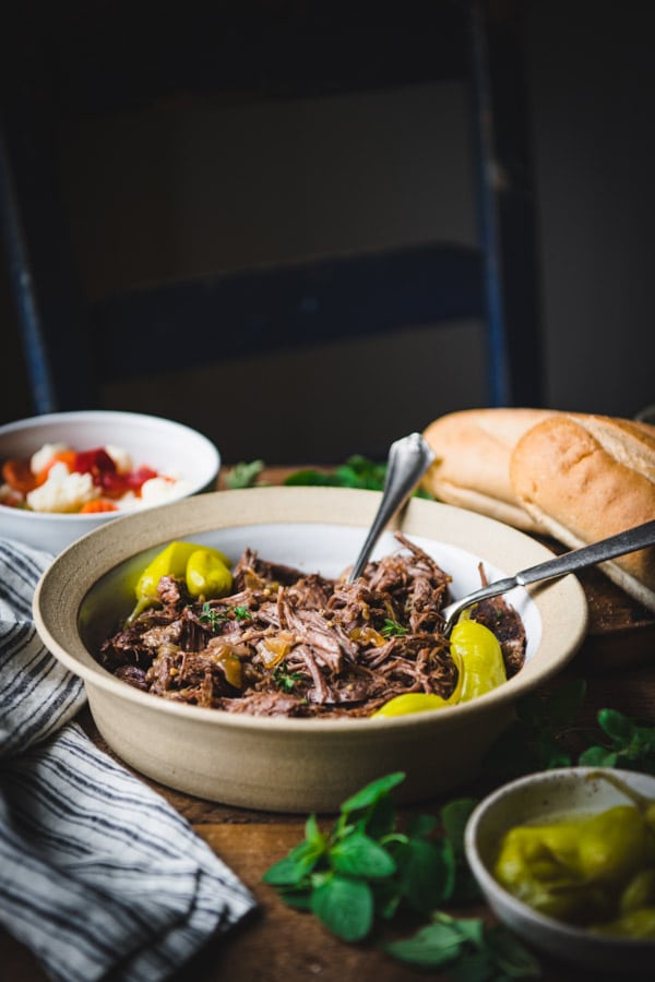 A bowl of shredded beef on a table, surrounded by the ingredients needed to make an Italian beef sandwich - hoagie rolls and a bowl of giardiniera.