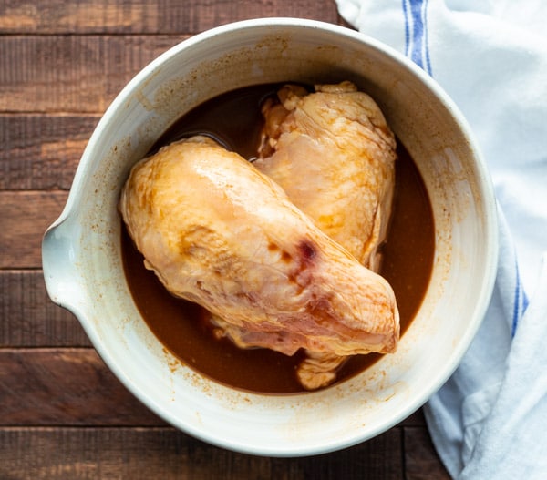 Two bone-in, skin-on chicken breasts marinate in a bowl of molasses and apple cider sauce.