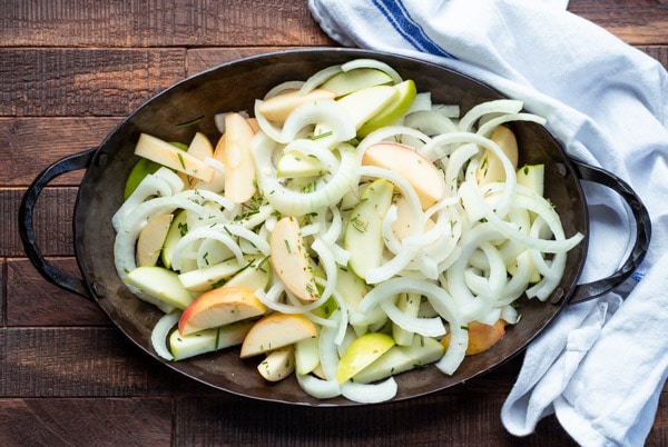 A roasting pan filled with raw sliced apples, onions, and fresh herbs.