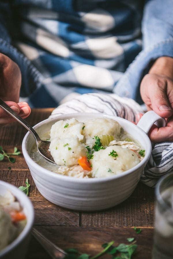 Hands eating a bowl of chicken and dumpling soup on a wooden farmhouse table