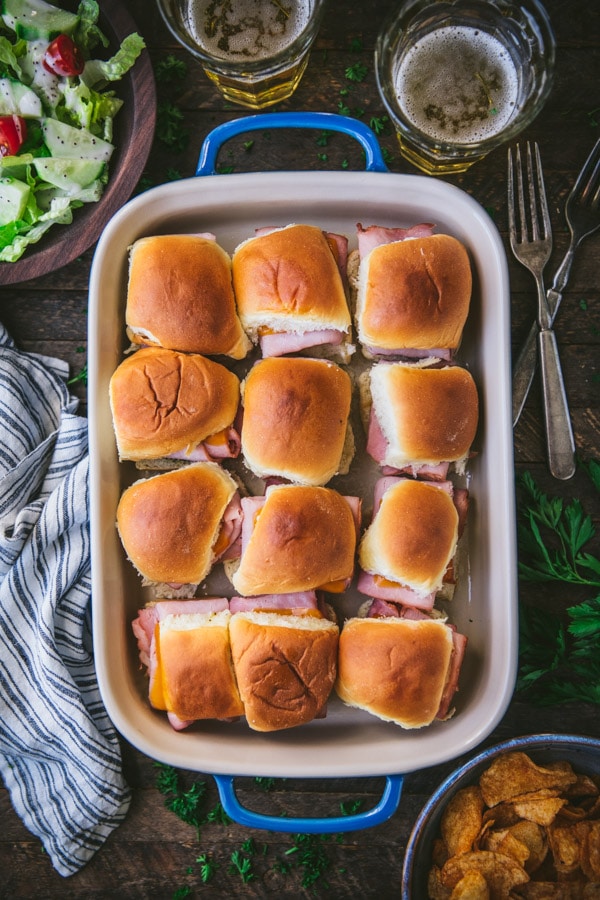 Hot ham and cheese sliders in a baking dish on a wooden table served with a side salad and potato chips