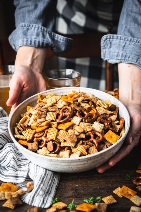 Hands serving a bowl of homemade Chex mix on a wooden table