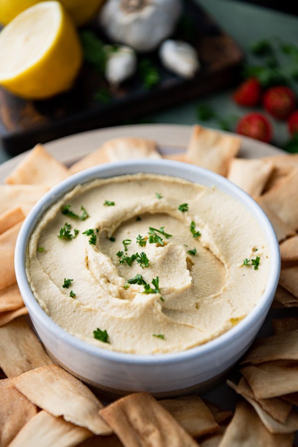 Close up side shot of a bowl of the best hummus recipe garnished with parsley and served with a plate of pita chips