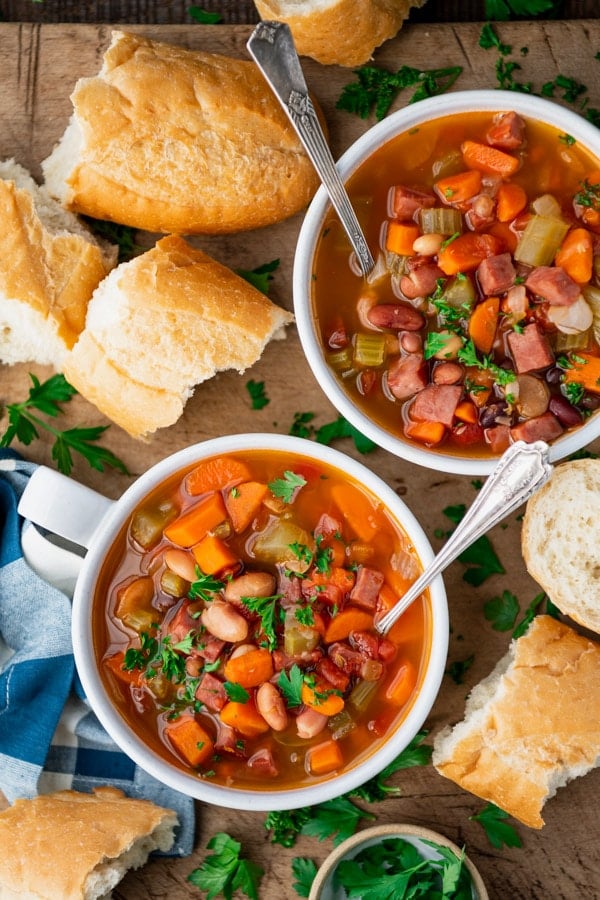 Overhead shot of two bowls of 15 bean soup on a wooden board with a side of bread