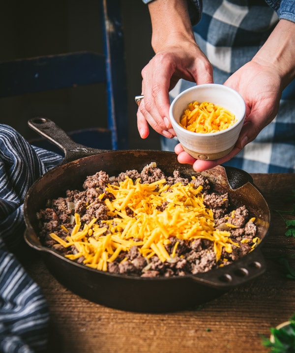 Adding shredded cheddar to a skillet of ground beef.