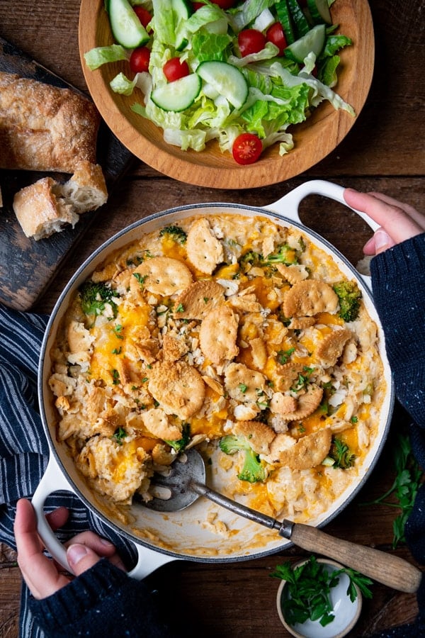 Hands holding a pan of chicken broccoli and rice casserole on a wooden table.