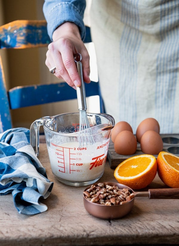 A women mixes the wet ingredients for cranberry bread in a glass measuring cup using a whisk. The cup sits on a tabletop alongside walnuts, an orange sliced in half, and four eggs.