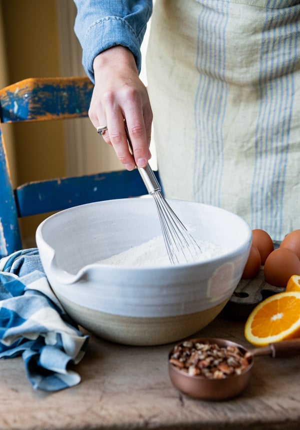 A woman whisks a bowl of flower. On the table next to the bowl is a measuring cup filled with walnuts, sliced oranges, and eggs.
