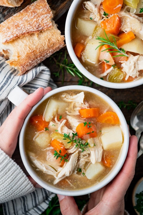 Hands holding a bowl of slow cooker chicken stew with a side of bread and fresh parsley garnish