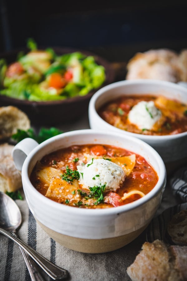 Two bowls lasagna soup on a table with ricotta topping and salad in the background