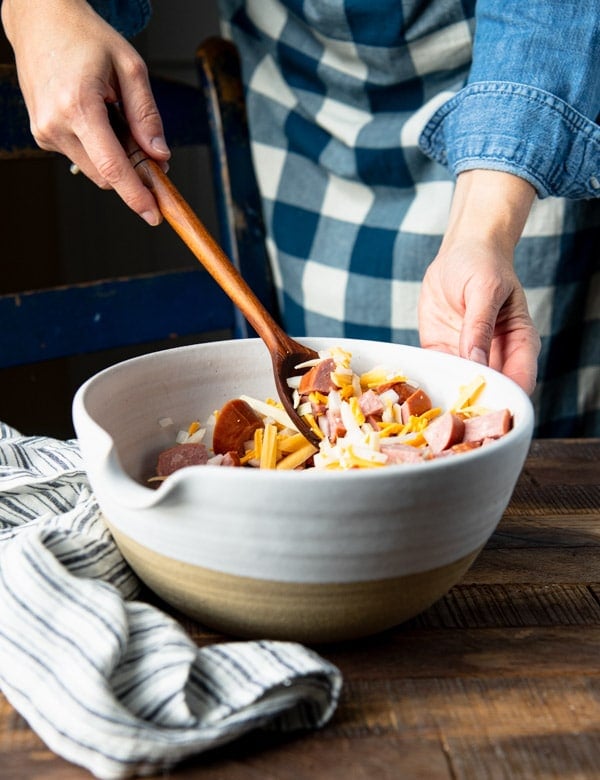 Stirring together kielbasa and potatoes with cheese in a white mixing bowl