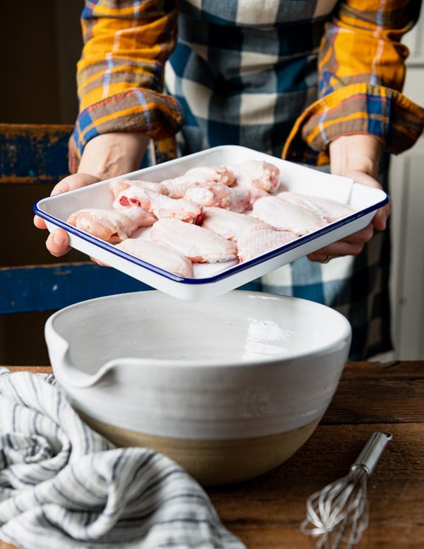 A woman holds a small tray full of raw bone-in, skin-on chicken wings over a large white ceramic bowl.