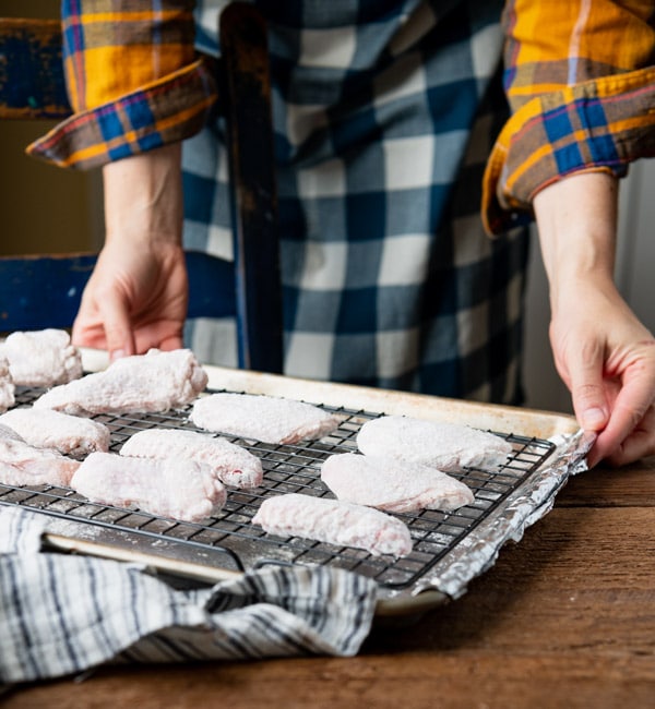 A woman prepares a foil-lined baking sheet for baking chicken wings. Flour-dredged wings sit on a wire rack, ready to be put in the oven.