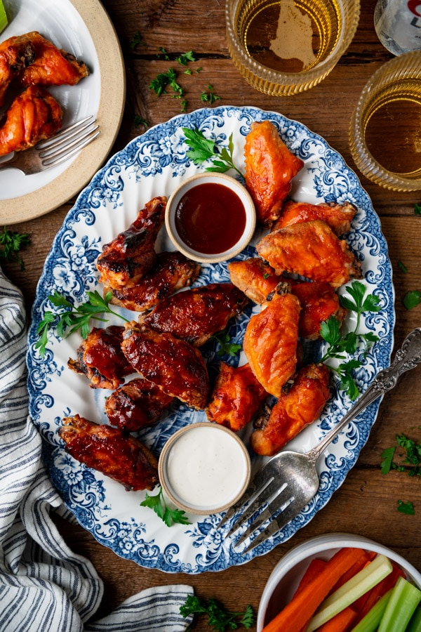 An overhead shot of a blue and white platter, filled with baked chicken wings. The wings are coated in BBQ sauce and buffalo sauce, and served with sides of ranch dressing and BBQ sauce for dipping.