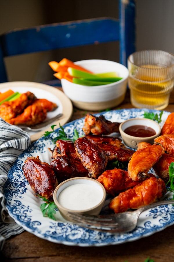 A platter of crispy baked chicken wings coated with BBQ sauce and buffalo sauce. The wings are served with ranch, BBQ sauce, and carrot and celery sticks as sides.