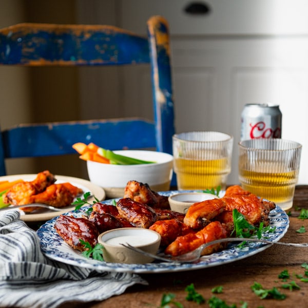 A platter of baked chicken wings served on a platter, alongside sides of ranch dressing, BBQ sauce, carrots and celery sticks, and two glasses of beer.