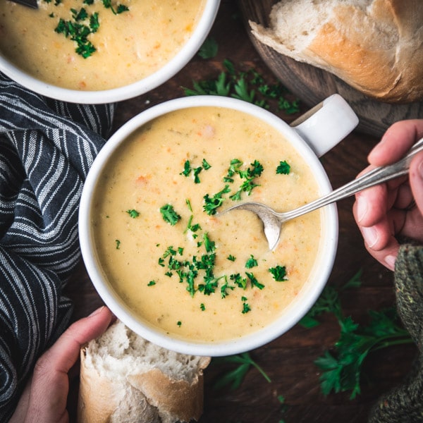 Hands holding a bowl of broccoli and cheese soup on a wooden table with a side of bread.
