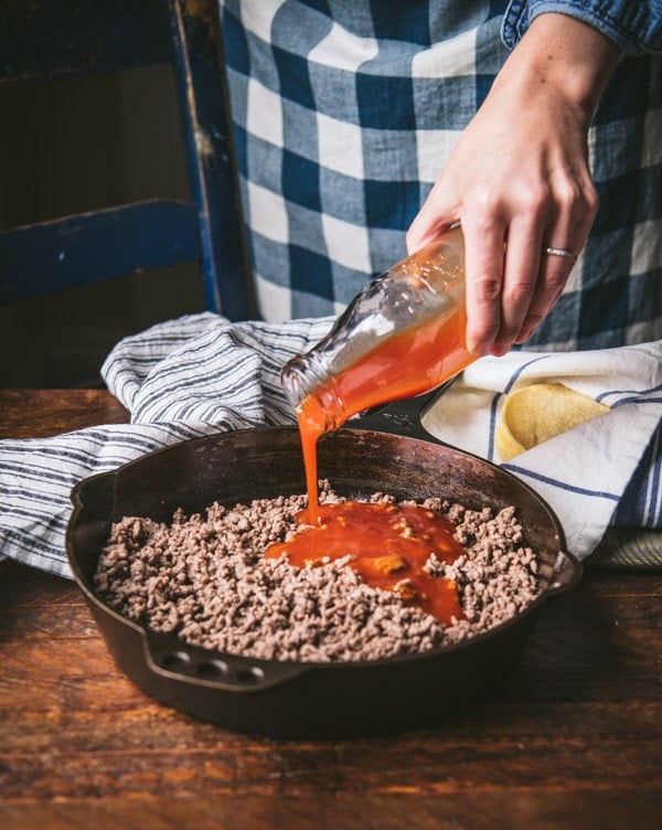 Pouring enchilada sauce into a skillet with ground beef.
