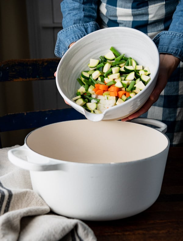 Adding a bowl of chopped vegetables to a white Dutch oven