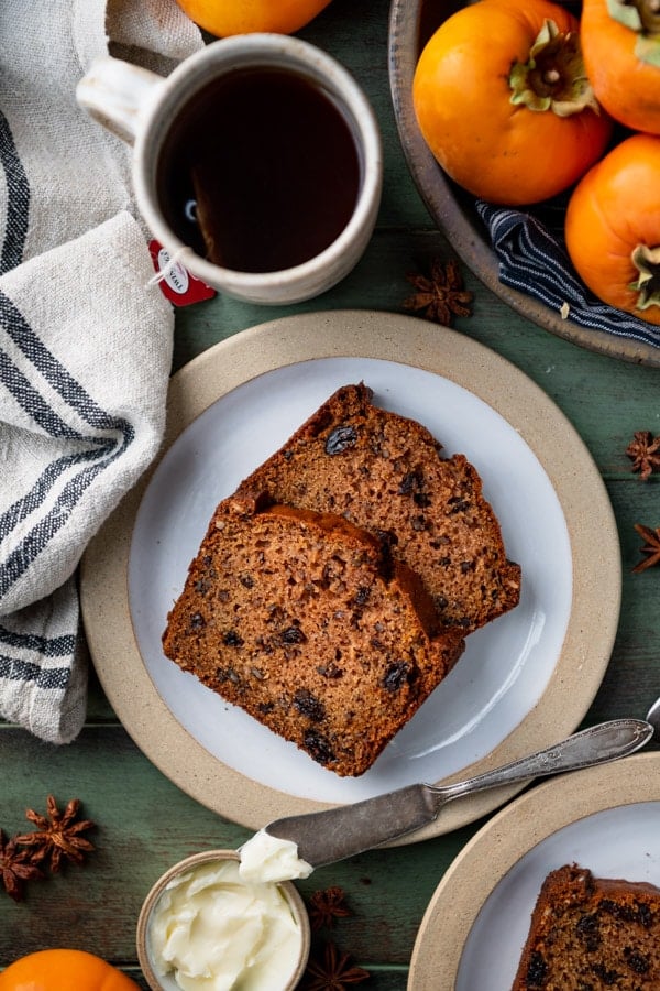 An overhead image of two slices of persimmon bread served on a plate with butter and a mug of tea.