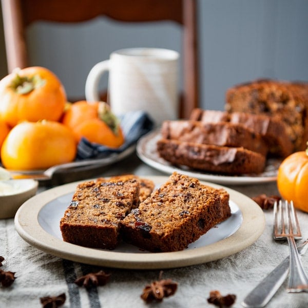 A square image of sliced persimmon bread served on a white ceramic plate. In the background are whole ripe persimmons and the full loaf of bread on a platter.