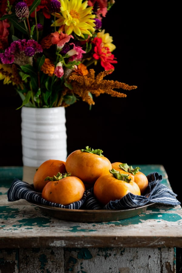 Side angle shot of a tray of five ripe orange persimmons stacked together. A case of colorful fresh flowers stands in the background.