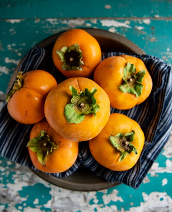 An overhead image of a bowl filled with five bright orange ripe persimmons.