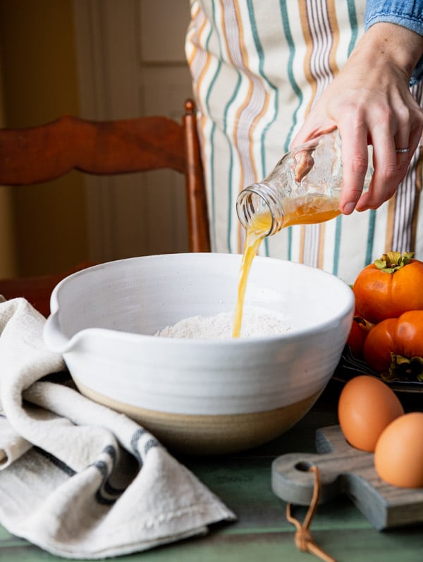 A woman pours a glass bottle of apple cider into a bowl containing dry ingredients for persimmon bread.