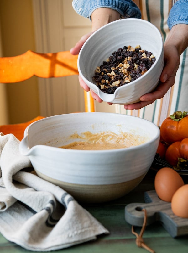 A woman pours a medium bowl of raisins and nuts into another larger bowl filled with persimmon bread batter.