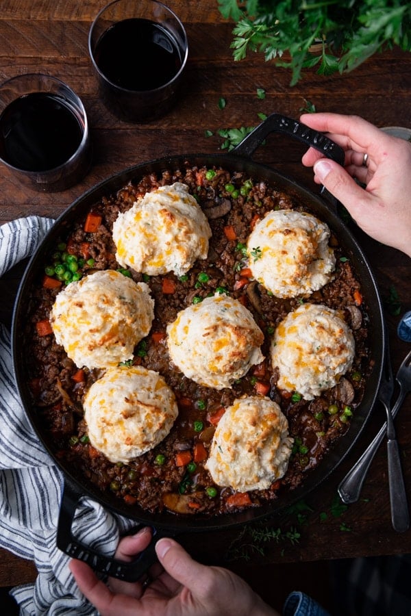 Overhead image of hands holding a cast iron skillet full of beef pot pie with cheddar biscuits on top.
