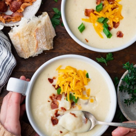 Overhead shot of hands holding a bowl of crockpot potato soup on a wooden dinner table.