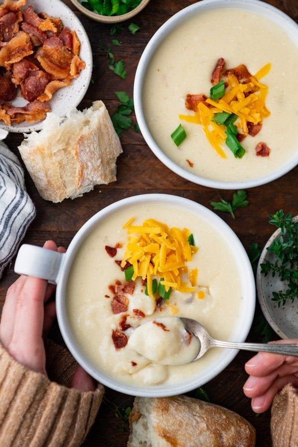 Overhead shot of hands holding a bowl of crockpot potato soup on a wooden dinner table.