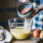 Adding ham to a glass mixing bowl full of eggs.