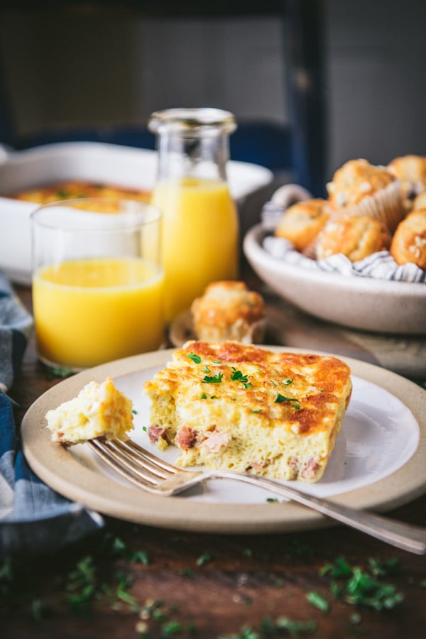 Side shot of a slice of baked ham and cheese omelette on a white plate with orange juice and muffins in the background.