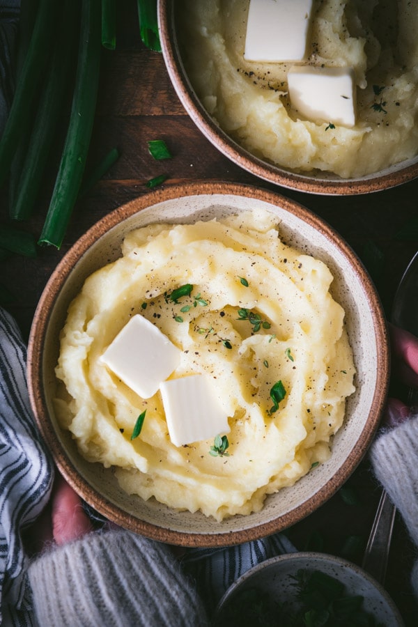 Overhead image of the best mashed potato recipe in a bowl on a wooden table with fresh herbs on top.