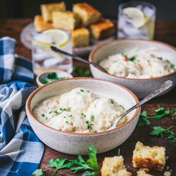 Two side by side bowls of chicken and dumplings in a creamy gravy. Each bowl has a fork and is served with cornbread.
