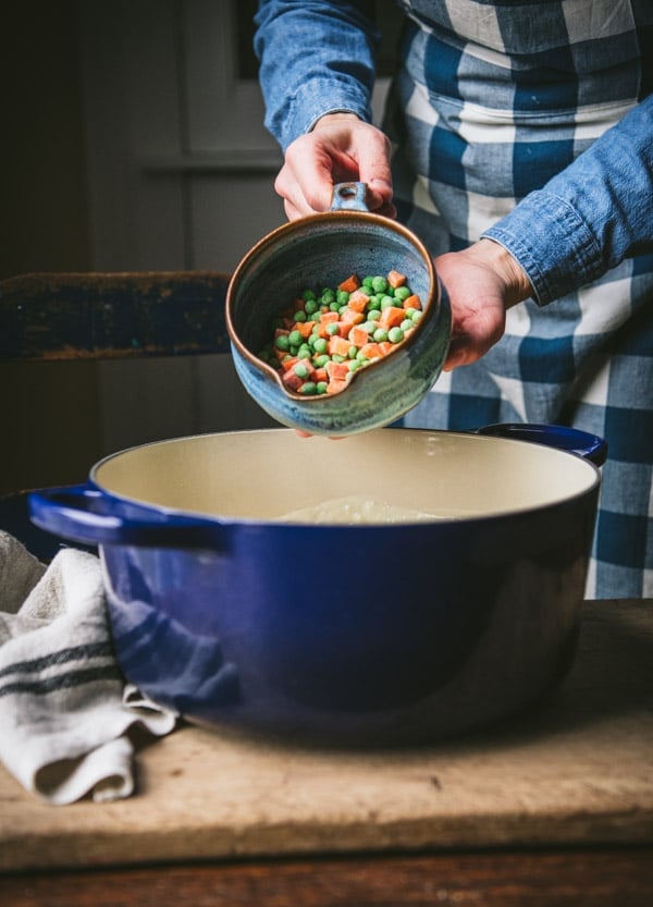 A woman pours a bowl of frozen chopped peas and carrots into a large blue enamel Dutch oven.