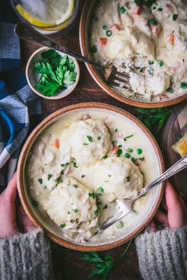 A woman holds a warm bowl of chicken and bisquick dumplings between her hands. Peas and carrots, shredded chicken, and fluffy dumplings sit in a creamy soup gravy, garnished with parsley.