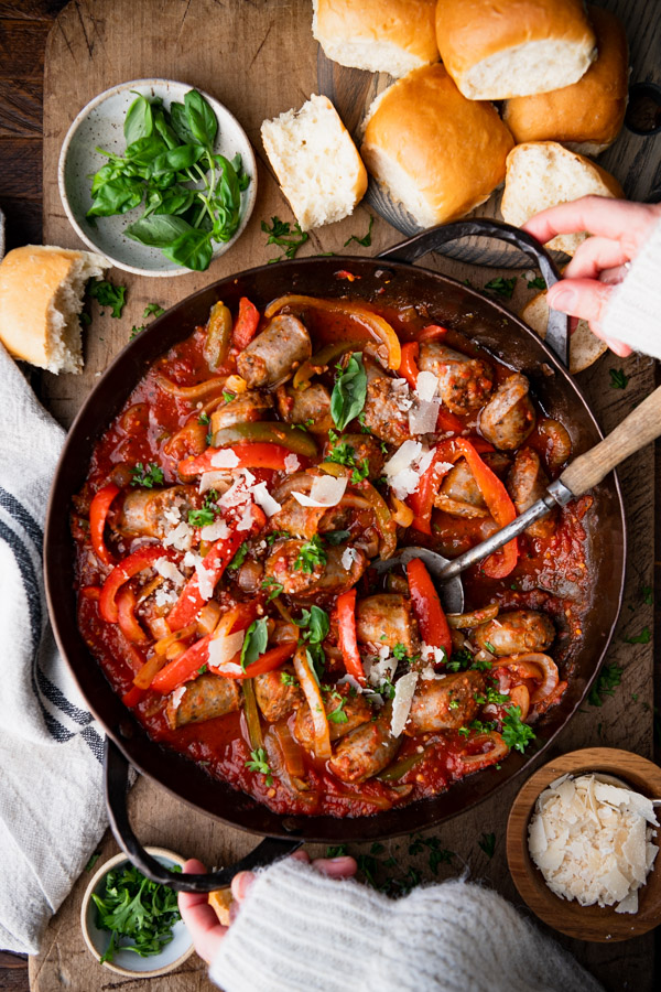 Hands holding a skillet of the best Italian sausage and peppers recipe on a wooden table with a side of bread
