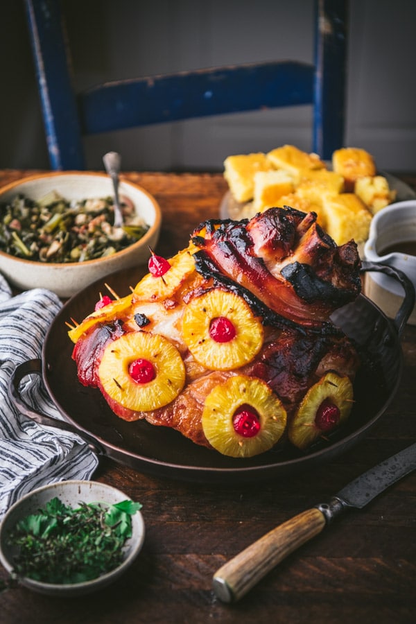 Pineapple and cherry ham in a roasting pan on a holiday table with collard greens and cornbread.