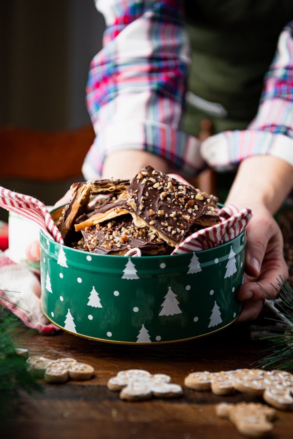 A woman places a tin of sweet and salty Christmas Crack toffee on a table. The tin is green with little white Christmas trees.