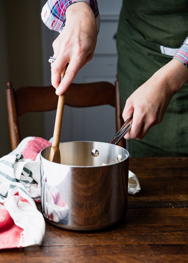 A woman stirs hot homemade toffee with a wooden spoon in a small metal saucepan.